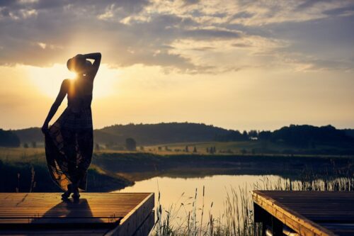 selbstbewusste Frau steht auf einem Steg am See und blickt in die Ferne zur Sonne