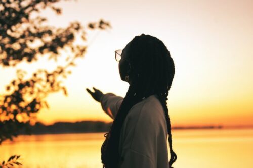 Frau steht am Seeufer und streckt ihre Hand Richtung Sonnenaufgang am Horizont